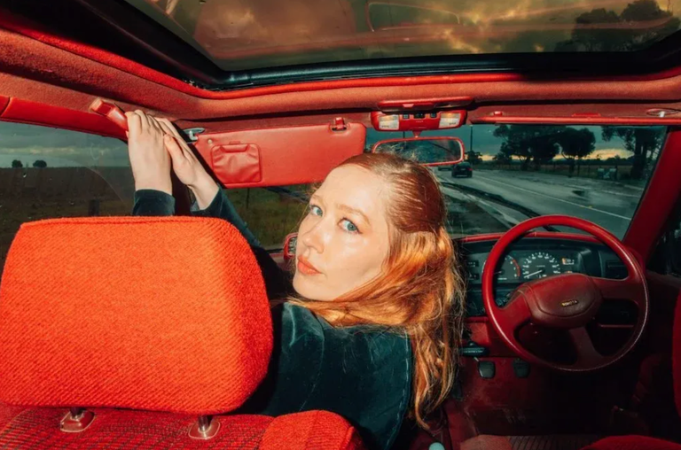 Australian singer-songwriter Julia Jacklin looking back from the front seat of a car with a vibrant red interior for a music promotional portrait.